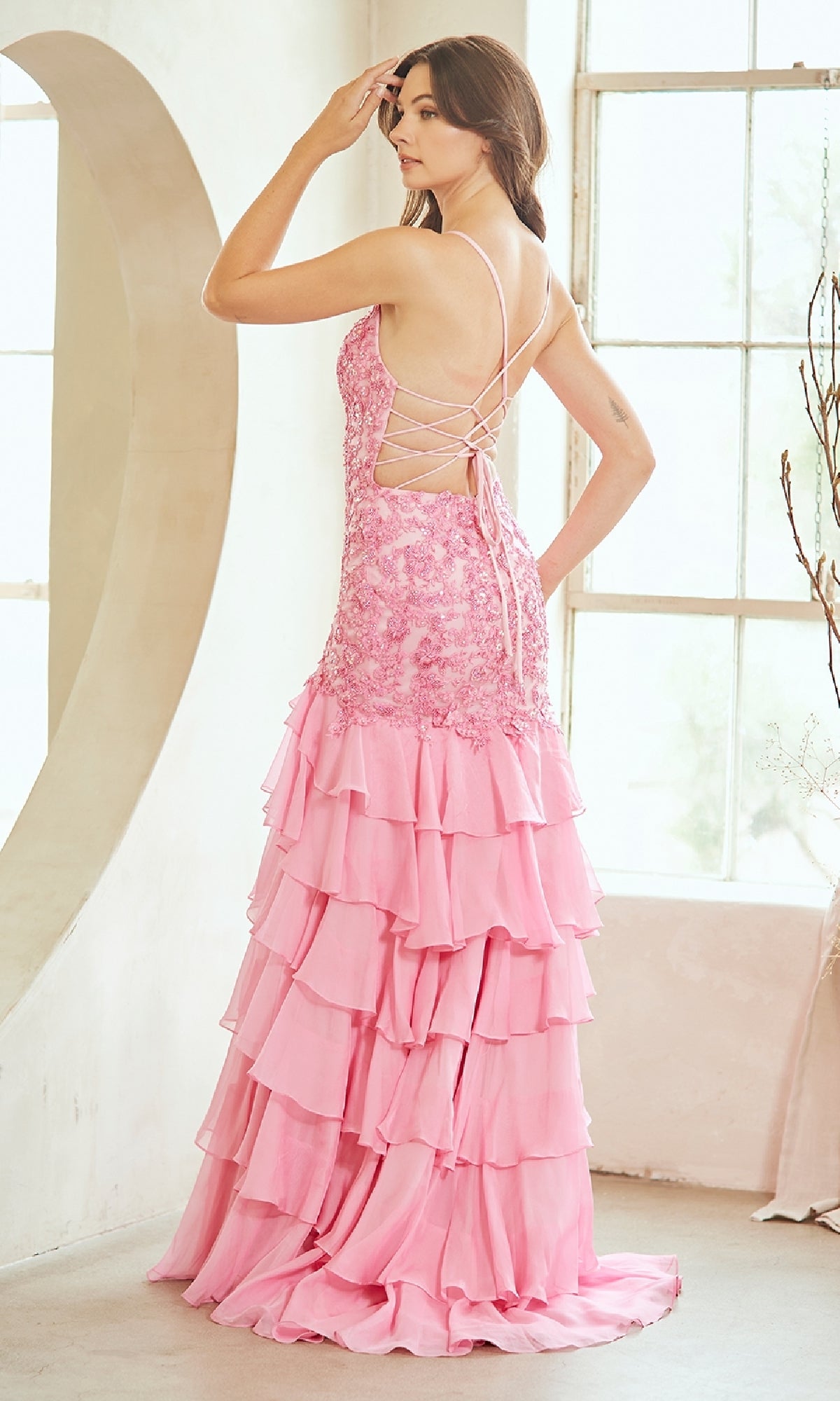 A woman stands indoors facing away from the camera, wearing the Amelia Couture 7085 Ruffled Long Prom Dress in pink with an embroidered bodice, lace-up back, and layered ruffles.